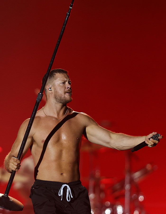 Dan Reynolds sin camisa en el escenario que contiene el soporte de micrófono, que actúa con fondo rojo relacionado con la masculinidad tóxica.