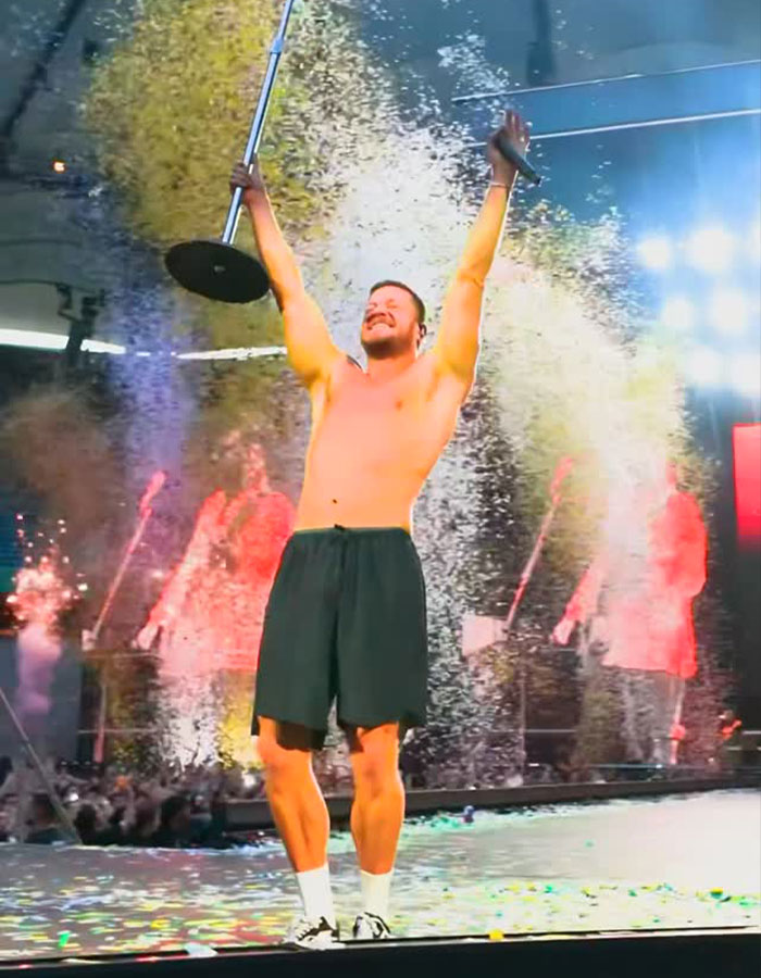 Dan Reynolds sin camisa en el escenario, sosteniendo el puesto de micrófono con confeti y luces que celebran el tema de la masculinidad tóxica.
