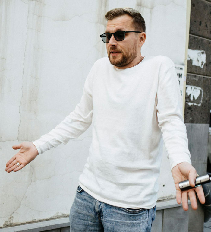 Hombre con gafas de sol y ropa casual encogiéndose de hombros afuera, expresando frustración por ser llamado en quiebra y usado como cajero automático.