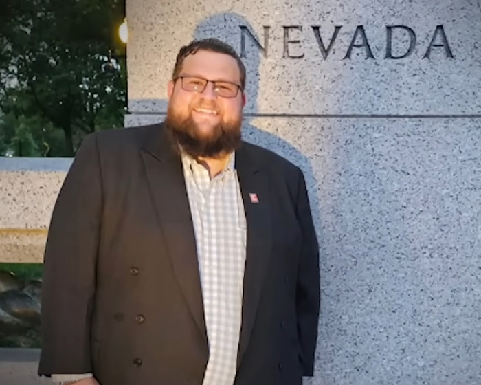 Hombre con gafas y barba sonriendo cerca de un monumento de Nevada, vinculado a la historia sobre acusaciones biológicas de padre y familia.