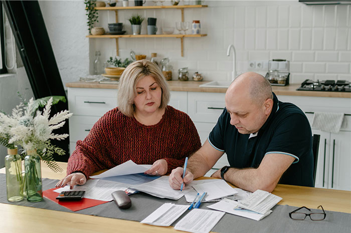 Padres de edad avanzada revisan las finanzas con documentos y calculadora, esperando que su hija apoye después de las pérdidas comerciales.