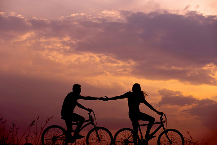 Silueta de una mujer y un hombre en bicicletas que se extienden entre sí durante el atardecer, simbolizando el conflicto de relaciones en línea.