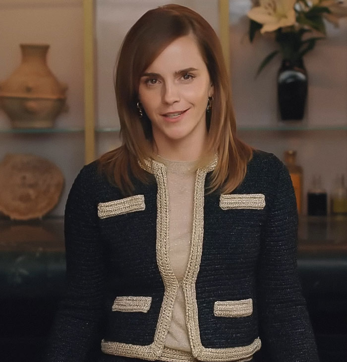 Emma Watson con un atuendo elegante en el interior, posando con confianza con un fondo natural y una sonrisa sutil.