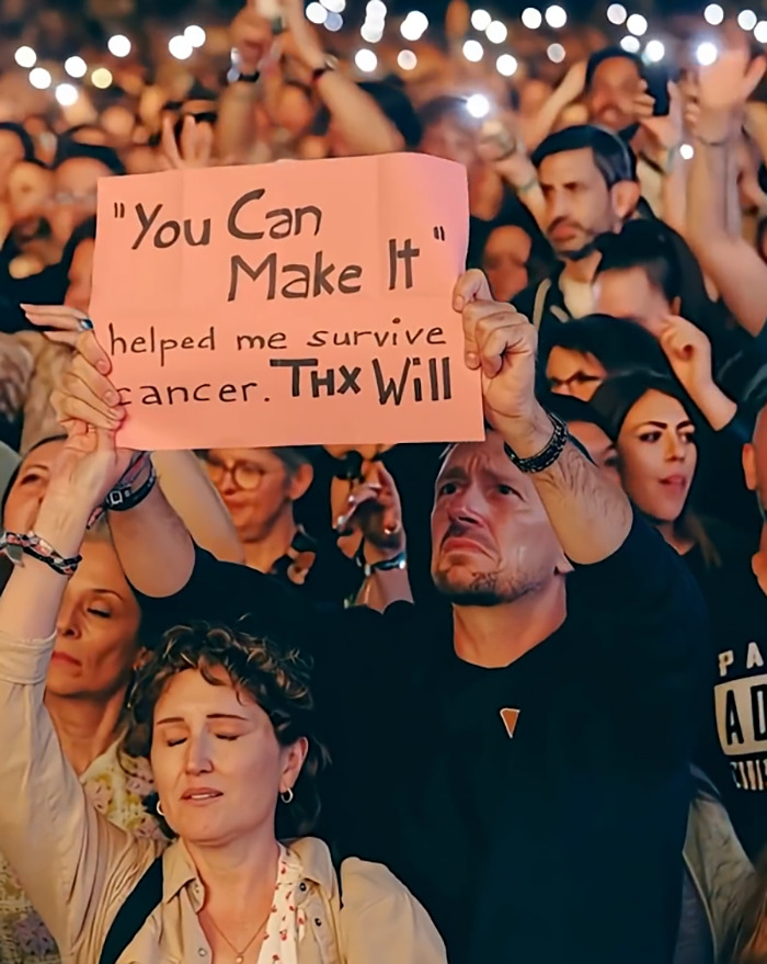 Multitud sosteniendo un letrero en un concierto, con muchas manos levantadas en el aire, destacando las discusiones de Will Smith y AI.