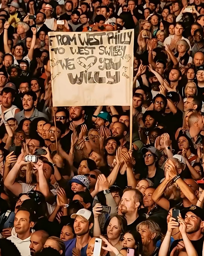 Multitud en el evento que tiene letrero para Willy con muchas manos levantadas en el aire, relacionadas con Will Smith fingiendo multitudes con IA.