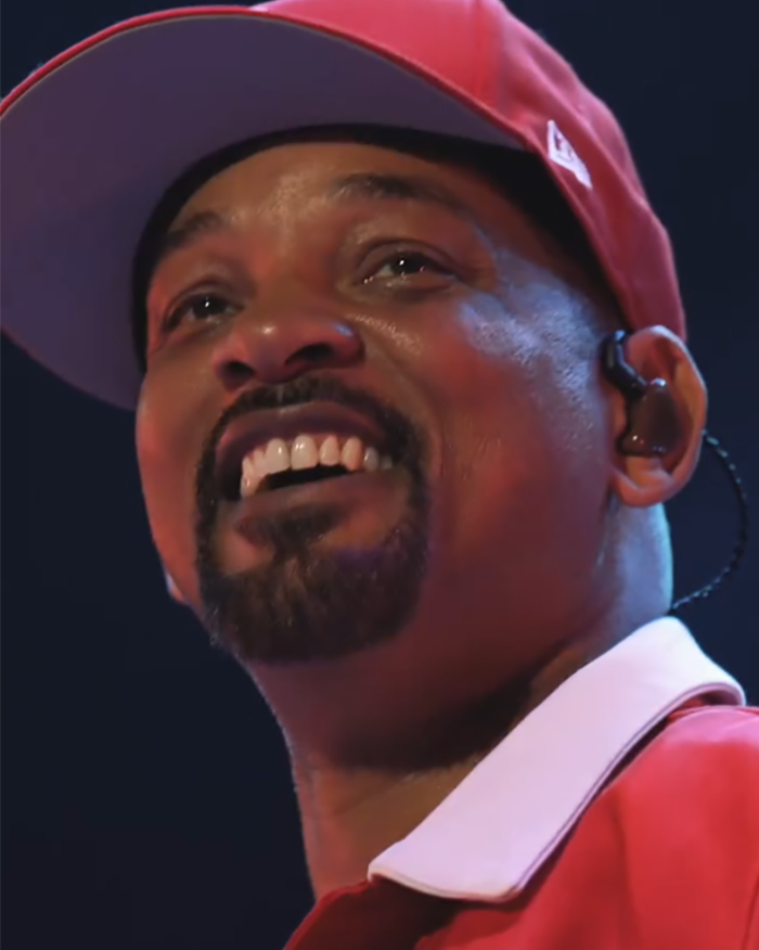Will Smith sonrió en el escenario con una gorra y una camisa roja, relacionada con ser burlado por fingir multitudes con IA.