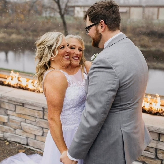 Gemelos Hensel en vestidos de novia tomados de la mano con novios al aire libre cerca del pozo de fuego, destacando la maternidad y el matrimonio.