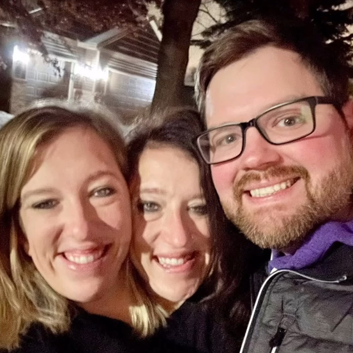 Tres adultos sonrientes al aire libre por la noche, relacionados con los gemelos Hensel New Baby y la especulación de la maternidad.