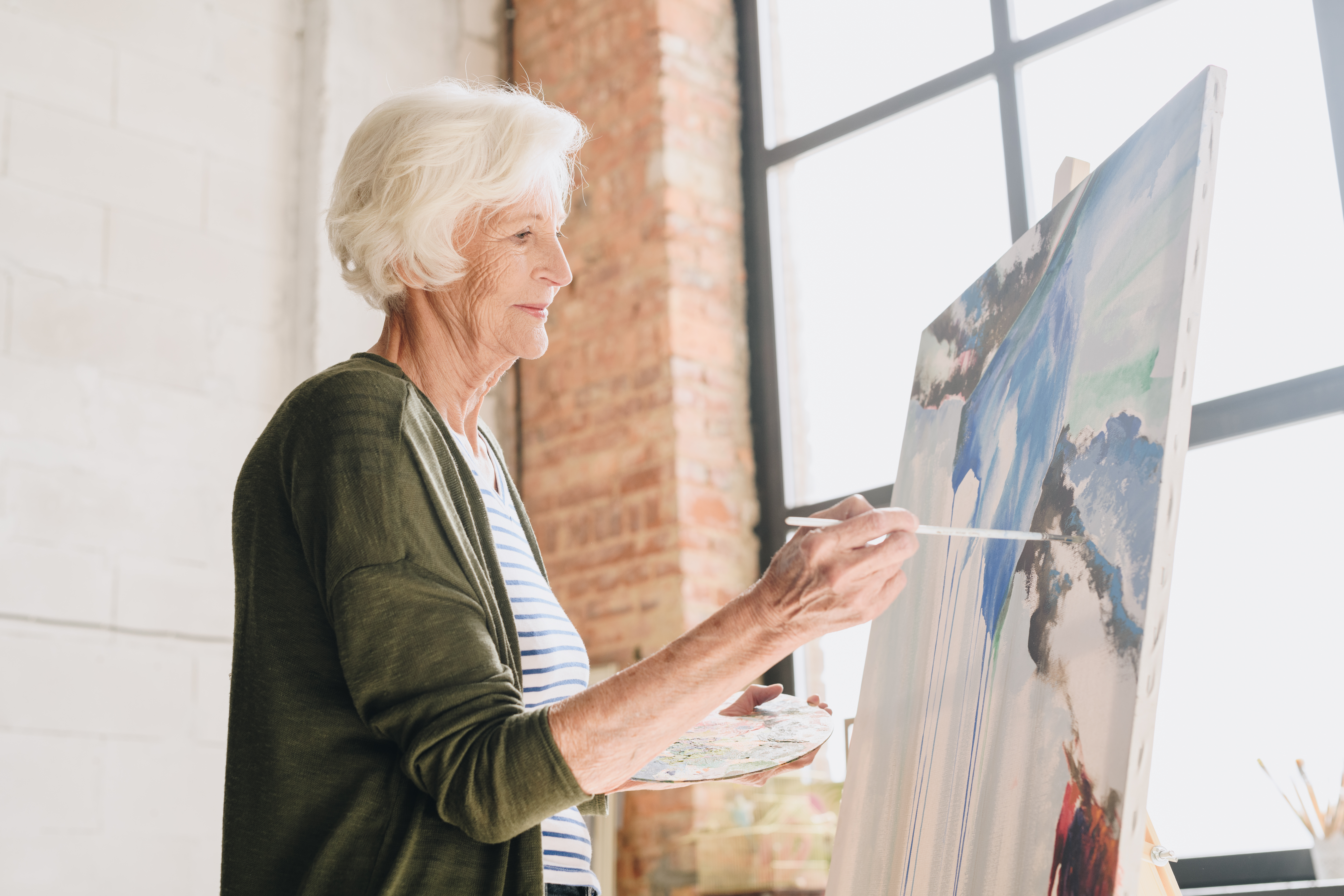 Mujer anciana pintando sobre lienzo en un estudio brillante, expresando creatividad e inspiración artística inesperada.