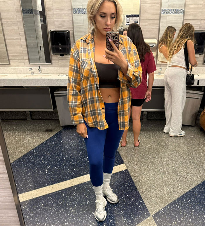 Modelo tomando selfie de espejo en leggings y franela abierta, destacando la discriminación de los senos grandes por controversia a la azafata.