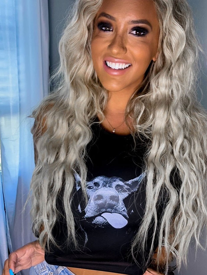 Modelo rubio con cabello largo y ondulado sonriendo, con una camisa negra, involucrada en un reclamo de discriminación de senos grandes.