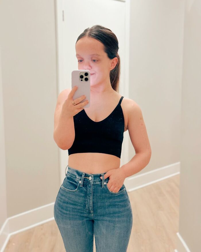 Mujer de 31 años con enanismo tomando un espejo selfie con top corto negro y jeans de cintura alta que muestran transformación después de la cirugía de altura.