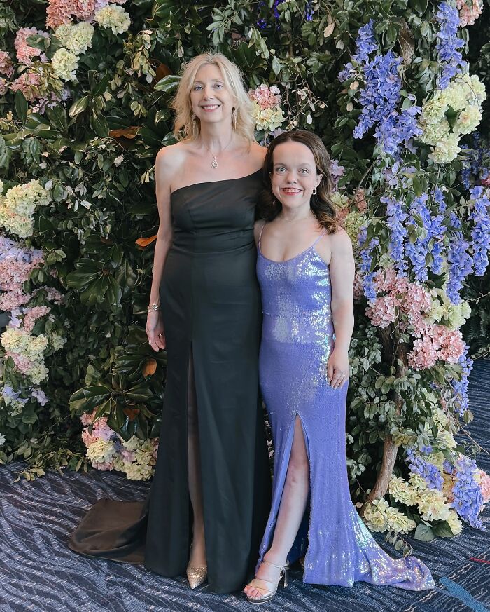 Mujer de 31 años con enanismo en un vestido morado brillante posando con una mujer más alta con un vestido negro contra un telón de fondo floral