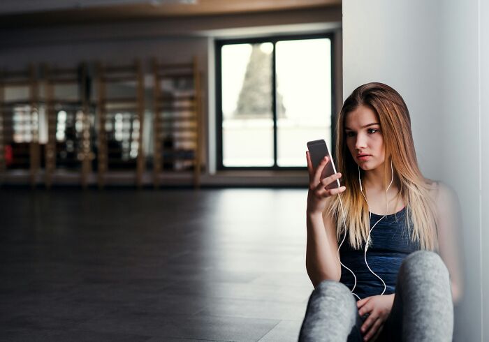 Mujer joven sentada sola en un gimnasio, mirando su teléfono, que ilustra a los clientes de la Generación Z comportamiento preocupante y salud mental.