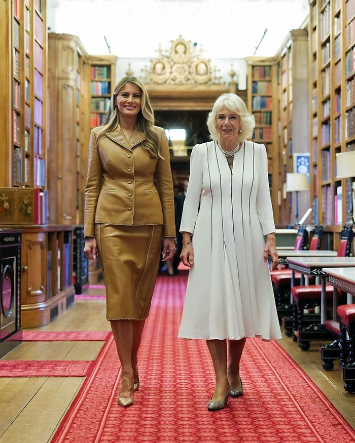Melania Trump con un traje de falda de cuero tostado junto a una mujer con un vestido blanco en una gran biblioteca del Reino Unido.