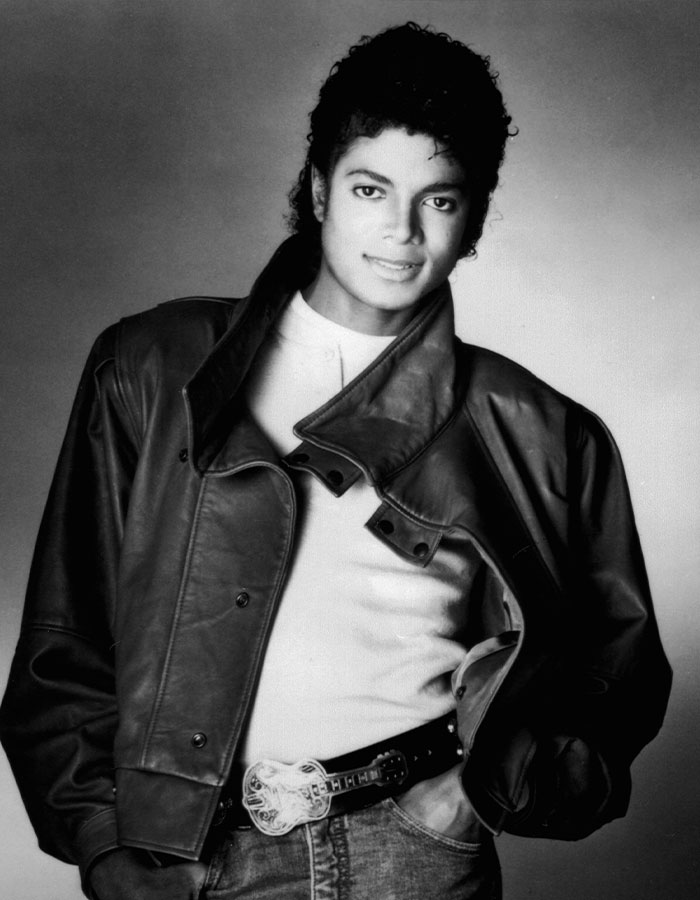 Michael Jackson con una chaqueta de cuero negra y una camisa blanca, posando con confianza con las manos en bolsillos en una clásica foto en blanco y negro.
