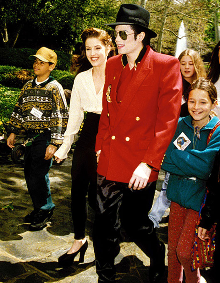 Michael Jackson con chaqueta roja y sombrero caminando al aire libre con un grupo sonriente, relacionado con las revelaciones de la vida íntima de Priscilla Presley.