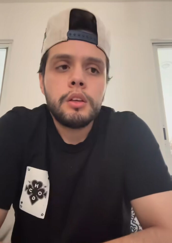 Young Man con una camisa negra y una gorra hacia atrás, pareciendo reflexivo en un entorno interior brillante.