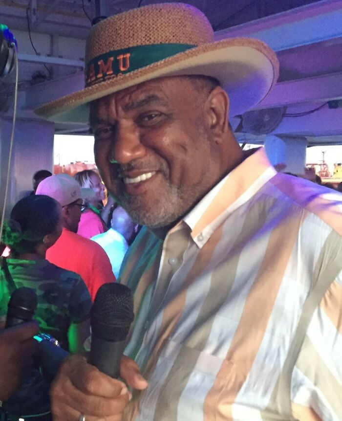 Hombre con un sombrero de paja sosteniendo un micrófono, sonriendo en un evento con una multitud en el fondo durante el rendimiento del medio tiempo.