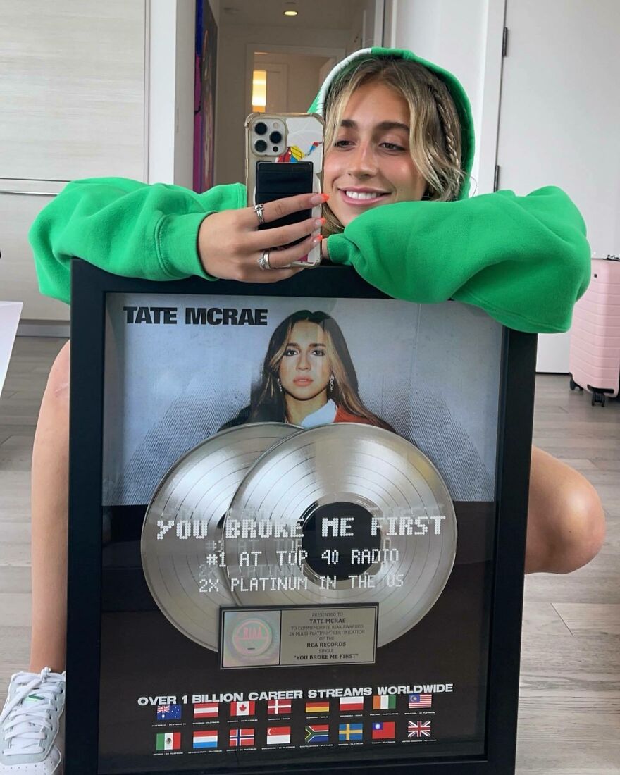 Tate McRae en una sudadera con capucha verde que tiene un premio de disco de platino para ti primero me rompió, mostrando su éxito musical.