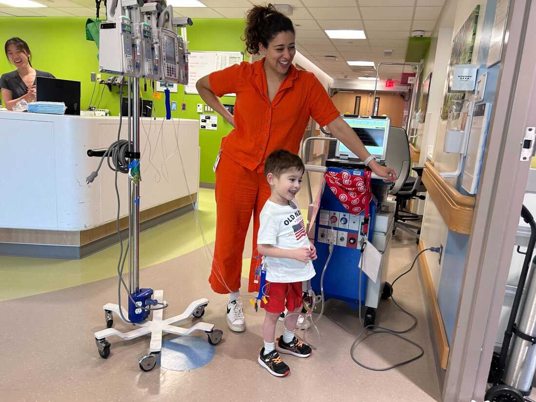 Caleb and his mother stand in a hallway at the hospital. Caleb is tethered to his ventricular assist device.
