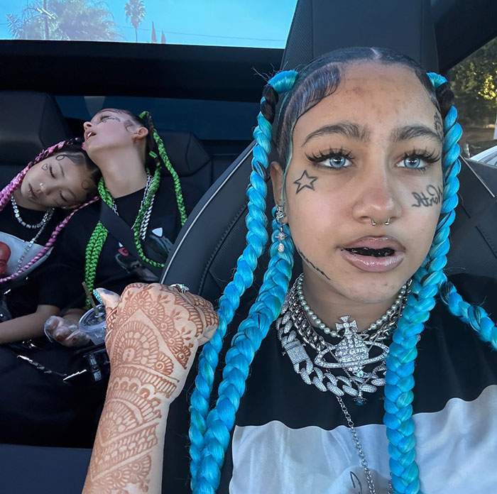North West con trenzas azules y tatuajes faciales sentada en un auto, destacando la advertencia de Kanye West sobre su vida en línea.