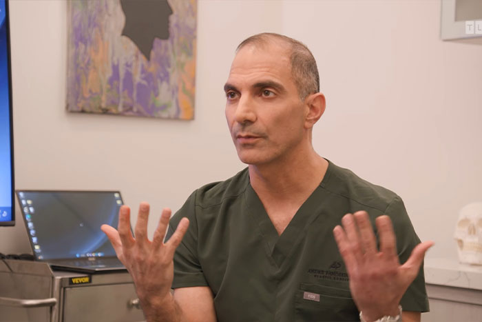 Médico con bata verde que explica inquietudes médicas urgentes sobre un hombre con un tumor facial de 4 libras en un entorno clínico.