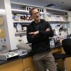 Timothy Koh is standing in his lab at the University of Illinois Chicago. Behind him are workstations with laboratory equipment and shelves with a variety of bottles and containers.