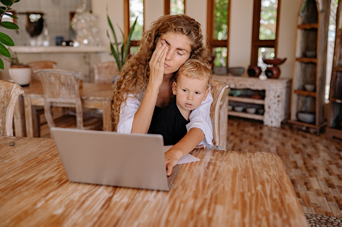 Mujer estresada sosteniendo a un niño, sentada en una mesa de madera con una computadora portátil, representando al hombre resentido con su esposa después del difícil tema del embarazo.