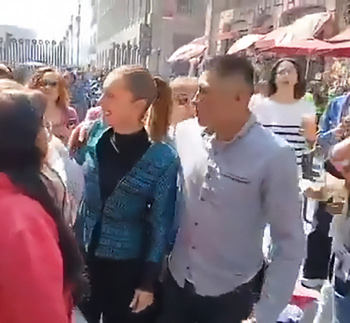 Hombre besando y acariciando al presidente mexicano mientras ella habla ante una multitud con gente y edificios en segundo plano.