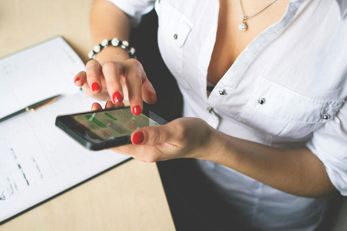 Persona con esmalte de uñas rojo usando un teléfono inteligente para charlar en familia sobre el dinero del cumpleaños, usando camisa blanca y pulsera plateada.