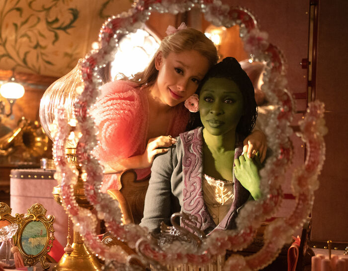 Cynthia Erivo y Ariana Grande se reflejan en un espejo decorativo durante una extraña escena de interacción.
