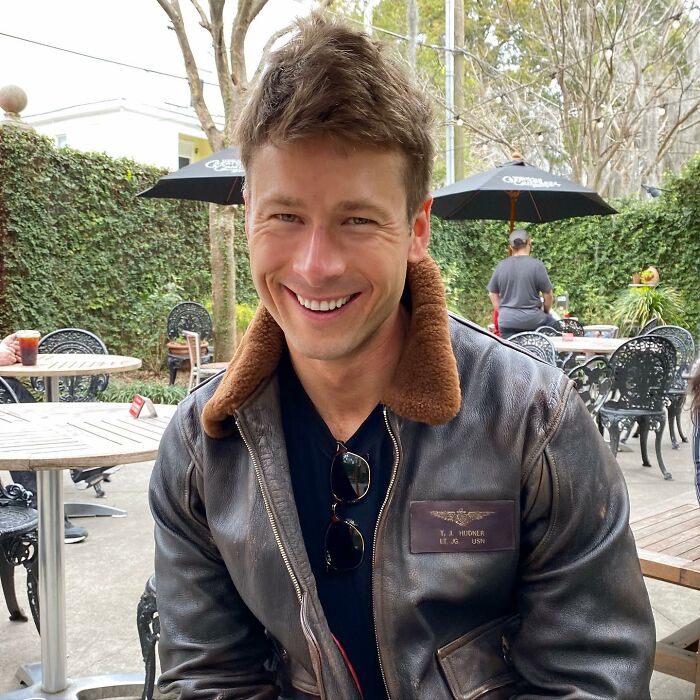 Hombre sonriente con una chaqueta de cuero marrón al aire libre, vinculado al error viral de Glen Powell y al lado oscuro de la discusión de Hollywood.