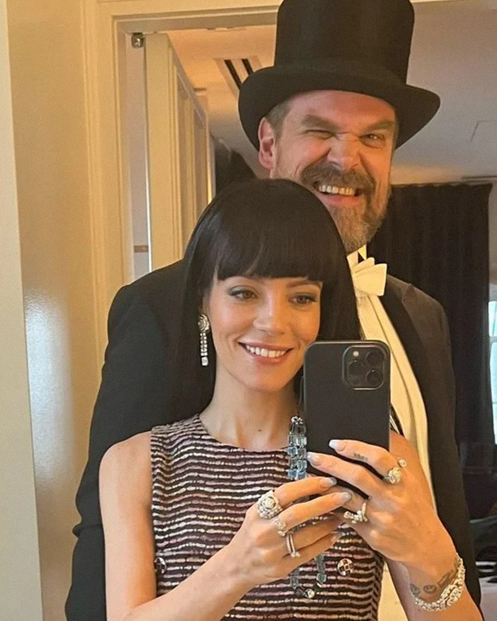 Alison Sudol sonriendo y tomándose una selfie en el espejo con David Harbour vestida con traje formal y sombrero de copa.