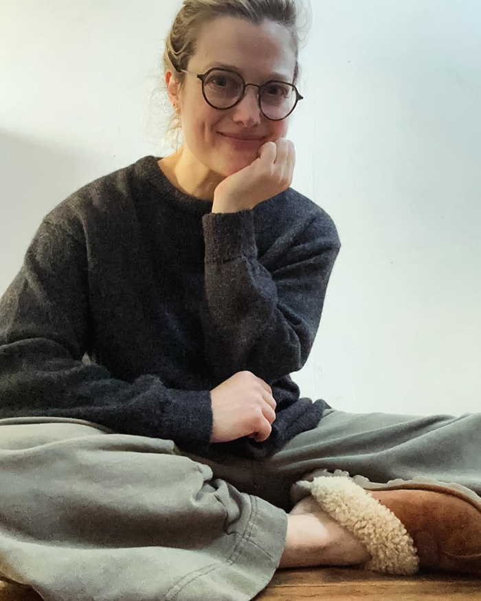 Alison Sudol sentada casualmente en el interior con gafas, un suéter oscuro y pantuflas cómodas, sonriendo gentilmente a la cámara.