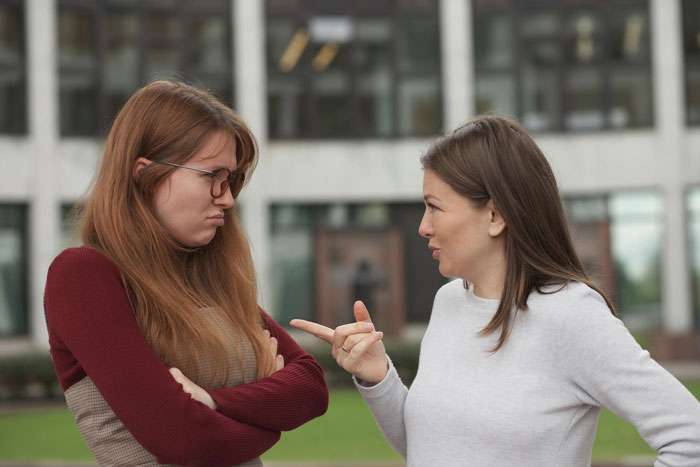 Mujer poliamorosa y novia en acalorado enfrentamiento fuera del edificio, expresando visiblemente frustración y tensión.