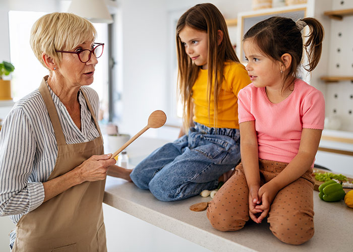 La abuela cuida a dos niños en la cocina, sostiene una cuchara de madera y les habla mientras se sientan en la encimera.