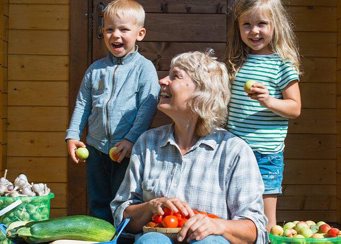 Abuela cuidando a cinco niños afuera, sosteniendo tomates y rodeada de verduras frescas en un día soleado.
