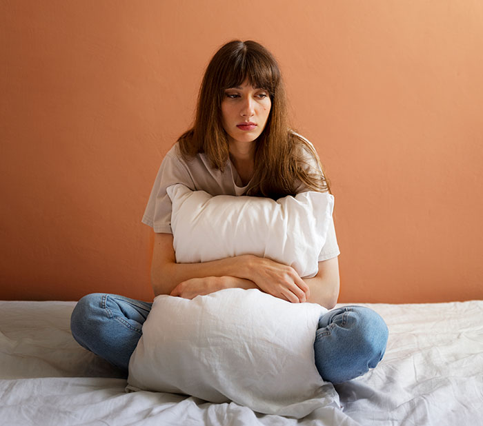 Mujer joven sentada en la cama abrazando una almohada, reflejando sentimientos heridos después de que un hombre corta los lazos con su madre por mensajes de texto a su novia