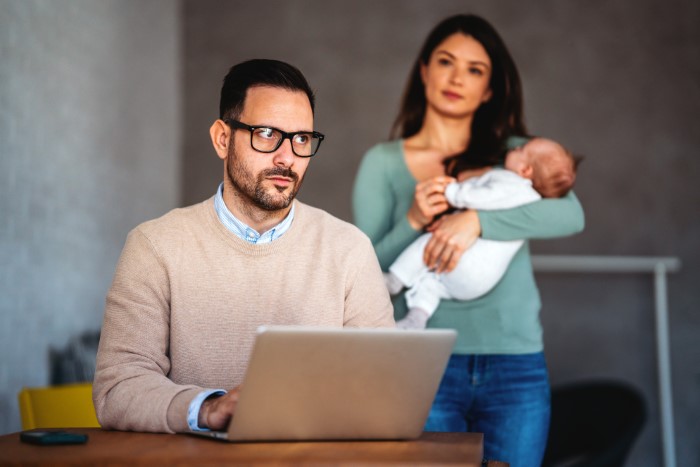 El hombre prioriza el trabajo en la computadora portátil mientras la esposa está detrás sosteniendo al bebé, mostrando distancia emocional en la familia.