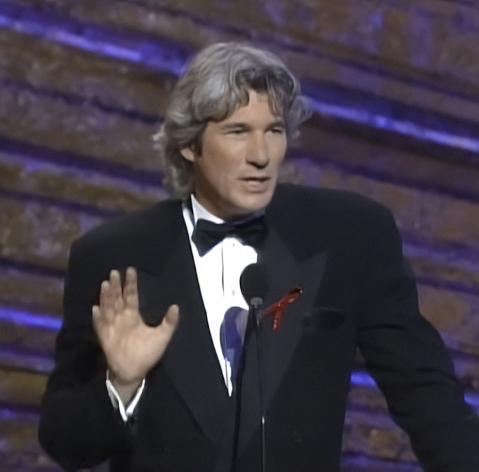 Richard Gere hablando en un evento, vestido con un esmoquin negro y dirigiéndose a la audiencia sobre la controversia sobre la prohibición de los Oscar.