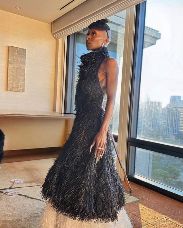 Cynthia Erivo posa en el interior con un vestido negro texturizado, resaltando sus manos extrañamente grandes a la luz natural junto a la ventana.
