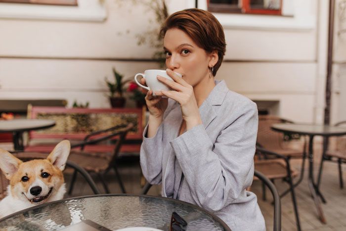 Mujer con una chaqueta ligera tomando café en una mesa de café al aire libre con un corgi feliz cerca, disfrutando de su cerveza asada oscura.