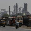 A motorcyclist and a three-wheel tuk-tuk pass a line of oil tanker trucks parked outside an oil refinery.