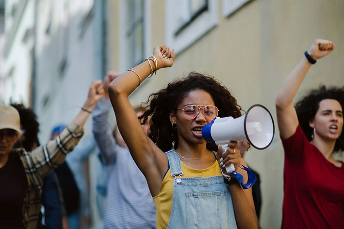 Una mujer con gafas y monos de mezclilla usa un megáfono encabezando una protesta contra los estándares de belleza con los puños levantados.