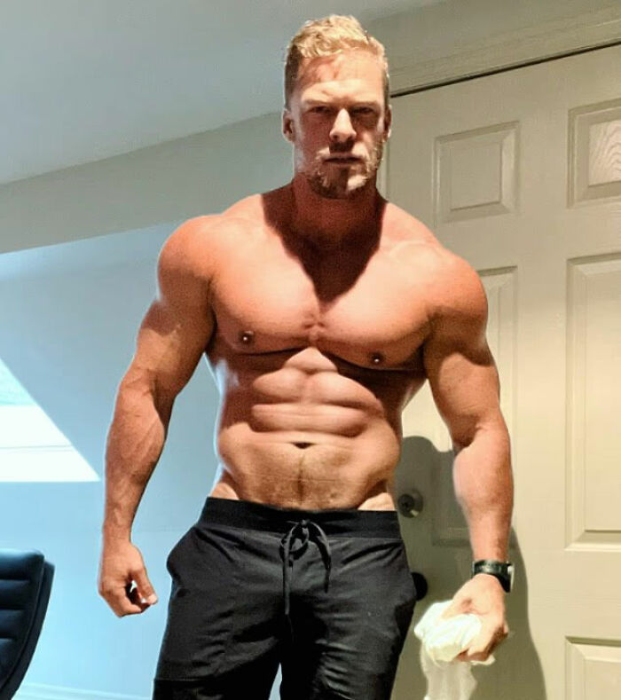 Hombre musculoso parado en el interior con pantalones cortos negros, relacionado con el vecino en pelea con la estrella de Reacher, Alan Ritchson.