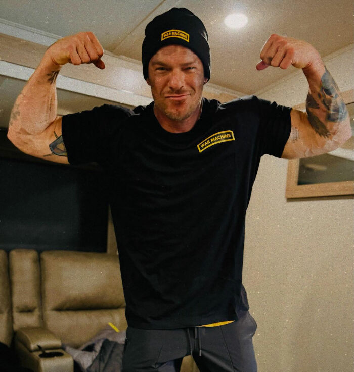 Un hombre vestido con un gorro y una camisa negros de War Machine flexionando los músculos en el interior en una disputa que involucra al vecino y estrella de Reacher, Alan Ritchson.