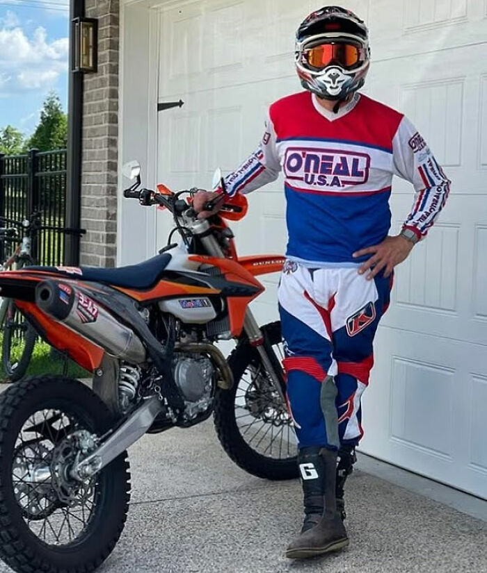 Hombre con equipo completo de motocross parado junto a una moto de cross fuera del garaje en una pelea vecina con la estrella de Reacher, Alan Ritchson