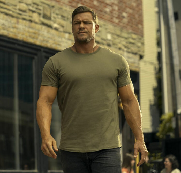 El actor Alan Ritchson, estrella de Reacher, caminando con confianza al aire libre con una camisa verde y pantalones oscuros durante un día soleado.