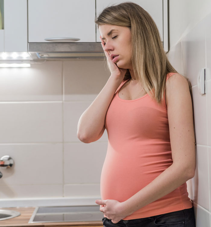 Mujer embarazada con un top rosa, sosteniendo su vientre y luciendo angustiada en una cocina moderna.